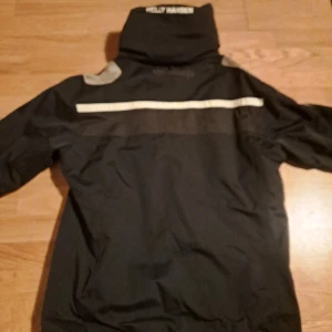Svart vindjacka från Helly Hansen - Svart vindjacka från Helly Hansen med vit HH-logga på bröstet och vita ränder över axlarna. Jackan har hög krage, dragkedja och praktiska fickor. Materialet är slitstark polyester och insidan har mjuk fleece. Perfekt för blåsiga dagar.
