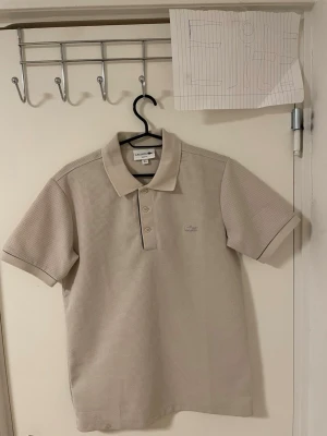 Beige piké Lacoste - Säljer en stilren beige pikétröja från Lacoste med klassisk krage och tre knappar framtill. Pikén har korta ärmar och diskret Lacoste-logga broderad på bröstet. Materialet är bomull med en lätt strukturerad yta och passformen är normal.Liknande modell som originala.