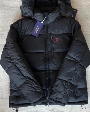 Svart pufferjacka från Polo Ralph Lauren - Svart pufferjacka från Polo Ralph Lauren med huva och tvåvägsdragkedja. Jackan har en röd broderad logga på bröstet och extra ficka med dragkedja. Materialet är polyester och jackan har en loose passform som är perfekt för kalla dagar. Storlek M pris 1500