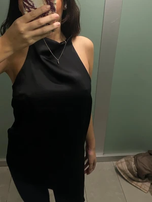 Svart ärmlös linnetopp i satin - Säljer en svart, slät och glansig linnetopp i satin med hög hals och ärmlös design. Toppen har en loose passform och ett elegant fall över kroppen. Perfekt att styla med jeans eller kjol för en trendig look.