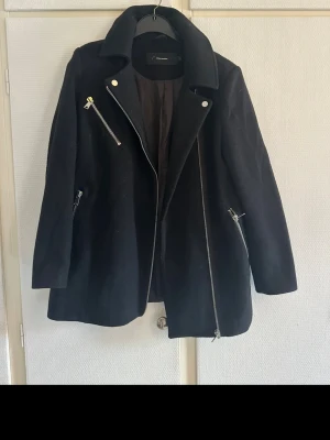 Svart ulljacka med dragkedjor Vero Moda - Svart jacka från Vero Moda i ullmix med coola silvriga dragkedjor och bred krage. Jackan har asymmetrisk frontzip och två snedställda fickor med dragkedja. Stilren och edgy look som passar perfekt till höst och vinter. Lite luddig och nopprig