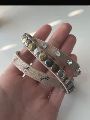 armband med nitar och stenar - Säljer ett beige rosa  armband med coola detaljer. Armbandet har både runda nitar i guld och silver samt glittrande stenar längs hela bandet. Det är designat för att vira flera varv runt handleden och har en snygg, lite rå känsla. Pris kan diskuteras 