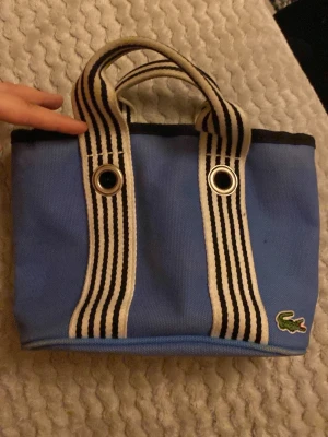 Blå handväska från Lacoste - Säljer en blå handväska från Lacoste med klassiska krokodilloggan. Väskan har vita och svarta randiga handtag, metallöglor och dragkedja med Lacoste-gravyr. Tillverkad i slitstark bomull, perfekt för att bära dina viktigaste grejer. Snygg och sportig design!