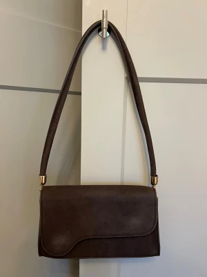 Brun mocka axelväska med gulddetaljer - Snygg brun axelväska i fuskmocka med stilrena gulddetaljer på axelremmen. Väskan har en mjuk, rektangulär form och magnetlås. Perfekt storlek för det viktigaste och har en enkel, modern look som passar till många outfits.