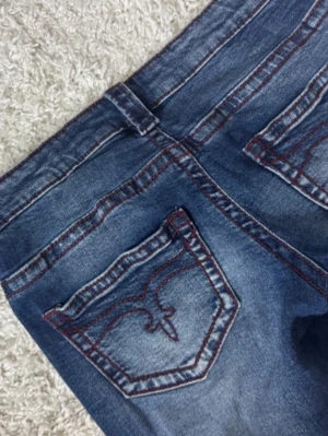 Blå bootcut jeans  - Säljer ett par snygga blå jeans med bootcut passform och låg midja. Jeansen har klassiska fickor med broderad detalj på baksidan och är tillverkade i jeansmaterial. Perfekt för dig som gillar retrovibbar och vill ha ett par jeans med lite extra attityd. Storlek 170=xs/s