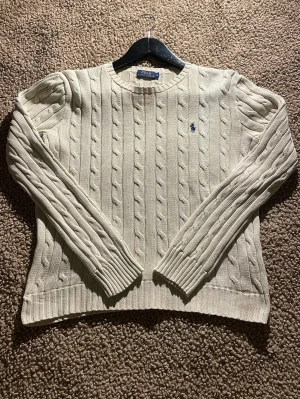 Beige kabelstickad tröja Polo Ralph Lauren - Snygg beige kabelstickad tröja från Polo Ralph Lauren med klassisk blå broderad logga på bröstet. Tröjan har rund hals, ribbade muddar och lång ärm. Perfekt för dig som gillar stilrena och tidlösa plagg med premiumkänsla.                                          !!!KRYMT I TVÄTTEN SÅ ÄR MER XS/S!!!