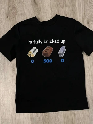 Svart t-shirt med rolig text - Svart t-shirt med trycket 'im fully bricked up' och grafik av stockar, tegelsten och metallbit med siffror under. T-shirten har rund hals och korta ärmar, perfekt för dig som gillar humor och memes. Materialet känns som mjuk bomull.
