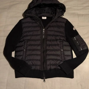 Moncler cardigan  - Svart pufferjacka från Moncler med huva och ribbade stickade ärmar. Jackan har dragkedja framtill och en ficka med dragkedja på ärmen där Moncler-loggan sitter. Materialet är en mix av polyester och stickat, perfekt för kyliga dagar.