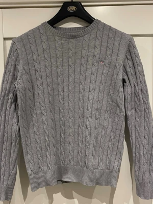 Grå kabelstickad tröja från GANT - Säljer en grå kabelstickad tröja från GANT i premium bomull. Tröjan har rund hals, ribbade muddar och ett litet broderat GANT-emblem på bröstet. Perfekt för dig som gillar klassisk och stilren design med snygga detaljer.
