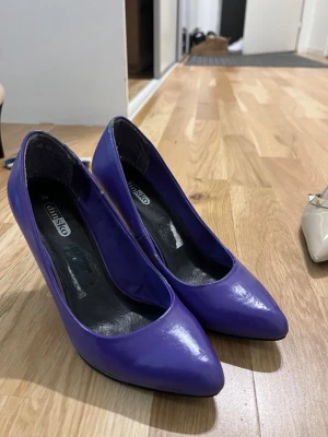 Lila pumps från DinSko med klack - Säljer ett par lila pumps från DinSko i storlek 36. Skorna har spetsig tå och hög klack, tillverkade i syntetmaterial med en blank finish. Perfekta för dig som vill sticka ut med färg på fötterna! 