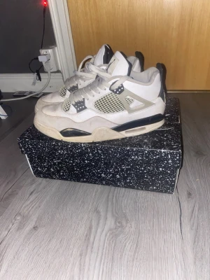 Nike Air Jordan 4 Retro vit/grå - Nike Air Jordan 4 Retro sneakers i vit, svart och neutralgrå färg. Klassisk meshpanel på sidorna, snörning och detaljer i plast. Snygg och ikonisk siluett med platt sula och rund tå. Perfekt för dig som gillar streetwear och basketstil.