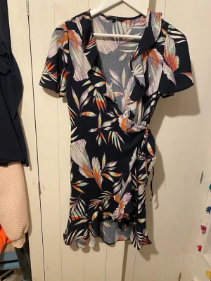 Blommig omlottklänning Vero Moda  - Snygg kort omlottklänning från Vero Moda i svart med färgglatt blommönster i lila, orange, grön och vit. Klänningen har volangkant, korta ärmar och knyt i midjan. Perfekt för sommarens alla tillfällen!