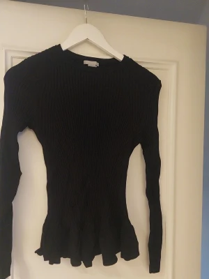 Svart ribbad stickad tröja med volang - Säljer en svart, ribbad stickad tröja med lång ärm och volangdetalj längst ner. Tröjan har en slim passform och är perfekt för dig som gillar en enkel men snygg look. Materialet är mjukt och stretchigt, vilket gör den skön att bära hela dagen.
