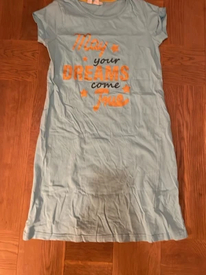 Ljusblå nattklänning från bpc - Ljusblå nattklänning från bpc bonprix collection med tryck i orange och svart framtill som säger 'May your dreams come true' och har små stjärnor. Väl använd. Nattklänningen är kortärmad och gjord i mjuk bomull, pyjamas. Pris kan diskuteras