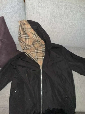 Svart Burberry vindjacka med huva - Svart vindjacka från Burberry med klassiskt rutigt foder i huvan. Jackan har dragkedja framtill, justerbar huva med snörning och två fickor. Materialet är lätt och känns som polyester, perfekt för blåsiga dagar. Stilen är clean och tidlös med Burberry London England-märke på insidan.