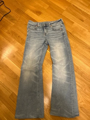Ljusblå bootcut jeans med fickor - Säljer ett par ljusblå jeans med bootcut-snitt och klassiska fem fickor, inklusive bakfickor med lock och knapp. Jeansen har en snygg tvätt med ljusa partier och är tillverkade i jeansmaterial. Perfekt för dig som gillar retrovibbar och bredare ben.