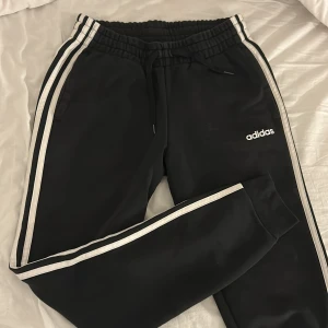 Svarta Adidas mjukisbyxor med vita ränder - Svarta mjukisbyxor från Adidas med klassiska vita ränder längs sidorna och Adidas-logga på benet. Byxorna har resår i midjan med snörning och är tillverkade i mjukt material som passar perfekt för chill eller träning. 