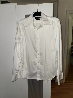 Vit skjorta från Brothers i bomull - Säljer en klassisk vit skjorta från Brothers i 100% egyptisk bomull. Skjortan har lång ärm, bred krage och knappar framtill. Perfekt för dig som gillar stilrena och enkla plagg med snygg passform. Extra knappar medföljer.