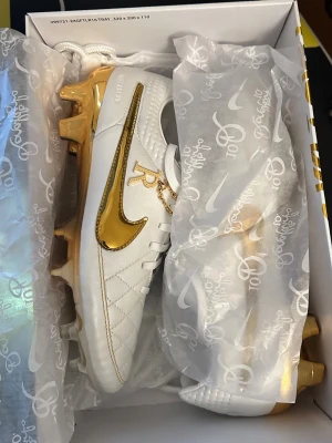 Nike Tiempo Legend 10R fotbollsskor - Storlek 42 Exklusiva Nike Tiempo Legend 10R fotbollsskor i vitt skinn med gulddetaljer och quiltad ovandel. Skorna har guldfärgad sula, snörning och en kedja med R-logga. Perfekta för dig som vill sticka ut på planen med stilrena och lyxiga detaljer.