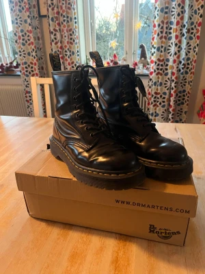 Svarta Dr. Martens boots med snörning - Ett par klassiska svarta Dr. Martens boots med grov platt sula och rund tå. Skorna har snörning och är tillverkade i blankt skinn med den ikoniska gula sömmen runt sulan. Skorna är änvända och har några små ytliga repor, inget som är särkilt synligt eller påverkar helhetsintrycket. Ett av snörena har en liten slitning på plaständen (aglet). Skriv gärna om fler bilder eller frågor 🌸🌸