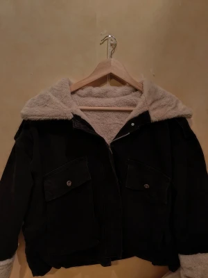 Svart teddyjacka med fluffig krage - Svart teddyjacka med stor fluffig beige krage och foder. Jackan har två stora fickor med knappar framtill och dragkedja. Materialet är jeans på utsidan och mjuk teddy på insidan, perfekt för kalla dagar. Ärmarna har också teddyfoder som syns vid mudden. S/M