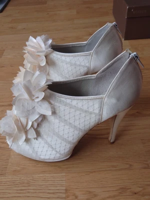 Vita pumps med blommor från Menbur storlek 40 - Säljer ett par festliga pumps från Menbur med öppen tå och hög klack. Klackhöjd ca 11,5cm främre platå ca 1,5cm. Färg Ivory/Gräddvit. Skorna har vackra blommor i tyg framtill, spetsdetaljer och dragkedja bak. Skorna har använts en gång så undersidan av sulan är det enda tecknet på användning vänligen se bilderna för referens.Skickas i originalkartongen för bästa förvaring. Skorna går ej att köpa längre då kollektionen utgått.