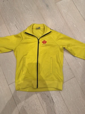 Gul ziptröja från Dsquared2 - Säljer en gul ziptröja från Dsquared2 med svart dragkedja och röd lönnlogga på bröstet. Tröjan har långärmade ärmar och svarta band med vita lönnblad längs axlarna. Orginal ( äkta )