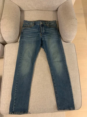 Blå jeans med slim fit - Snygga blå jeans med klassisk slim fit och fem fickor. Jeansen har en lätt tvättad look med subtila slitningar på låren och knäna. Tillverkade i robust denim med normal passform och midwaist. Perfekta för dig som gillar enkel och tidlös stil. Storlek 32/32