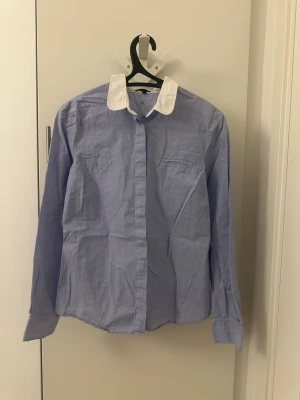 Blå skjorta med vit krage från Stockholm - Blå skjorta med kontrasterande vit krage och vita knappar. Klassisk rak passform och långärmad design. Tillverkad i bomull som känns lätt och fräsch. En bröstficka på vänster sida och stilrena manschetter med dubbla knappar. Perfekt för dig som gillar clean och enkel stil.