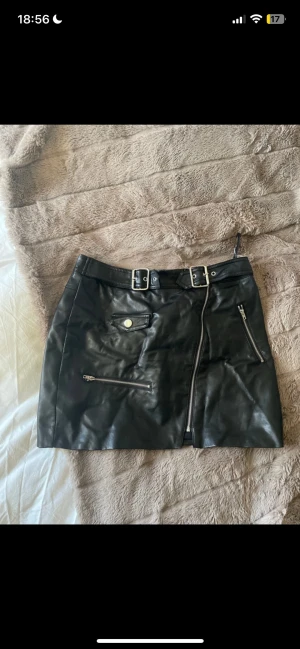 Svart skinnkjol från Forever 21 - Säljer en svart kort skinnkjol från Forever 21 med coola detaljer som dubbla bälten i midjan och flera dragkedjor framtill. Kjolen har fickor med knapp och en snygg, lite edgy look. Perfekt för dig som gillar statement-plagg och vill sticka ut. finns ett lite skav
