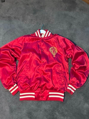 Vintage Röd bomberjacka  - Snygg röd bomberjacka med glansig finish och  luftballong print på bröstet. Jackan har vita och röda ränder på krage, muddar och nederkant, samt knappar framtill. Perfekt för dig som vill sticka ut med en cool retrostil.