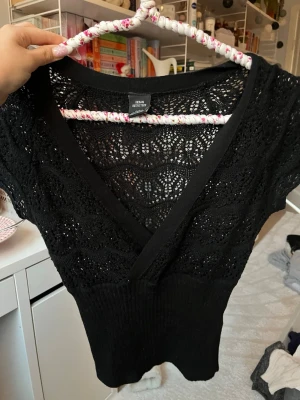 Svart spetsblus från Urban Outfitters - Supersnygg svart blus från Urban Outfitters med v-ringning och omlottkänsla. Toppen är stickad med spetsmönster och har korta ärmar. Perfekt att styla med jeans eller kjol för en trendig look.