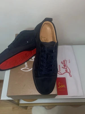 Svarta sneakers från Christian Louboutin - Snygga svarta sneakers från Christian Louboutin med klassisk röd sula och rund tå. Skorna har snörning och är tillverkade i mocka med diskreta metalldetaljer på sidan. Perfekta för dig som gillar exklusiv stil och vill sticka ut med lyxiga detaljer.