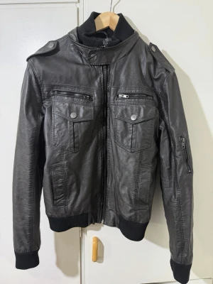 Svart skinnjacka från Indigo Jeans S - Snygg svart skinnjacka från Indigo Jeans i storlek S. Jackan har flera fickor med dragkedja och knappar, ribbade muddar vid ärmslut och midja samt coola axeldetaljer. Perfekt för dig som vill ha en edgy stil med streetvibe.