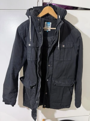 Svart parkajacka från WA Action Sports - Svart parkajacka från WA Action Sports med huva och snörning. Jackan har flera fickor med lock och knappar, samt dragkedja framtill. Materialet är slitstark polyester och jackan har en normal passform. Perfekt för kyliga dagar.