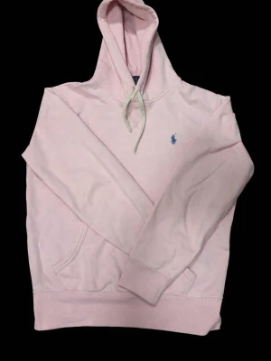 Ljusrosa Ralph Lauren Hoodie - Ljusrosa hoodie i mjukt och bekvämt material. Klassisk modell med huva, snören och känguruficka framtill. Broderad logotyp på bröstet. Perfekt för vardag och avslappnad stil. Bra skick.