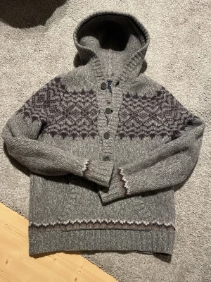 Grå stickad hoodie från Gap - Unikt stickad hoodie från Gap med snyggt mönster över bröstet och axlarna. Tröjan har huva, knappar framtill och en stor magficka. Perfekt för kalla dagar och har en klassisk nordisk vibe. Pris kan diskuteras.