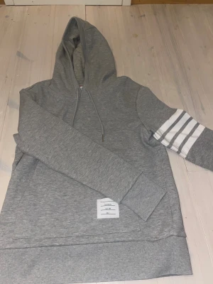 Grå hoodie från Thom Browne med ränder - Snygg grå hoodie från Thom Browne med huva och snörning. På ena ärmen finns fyra vita ränder som ger en sportig vibe. Tröjan har en vit patch med märkets logga framtill. Tillverkad i mjuk bomull, perfekt för chill dagar.