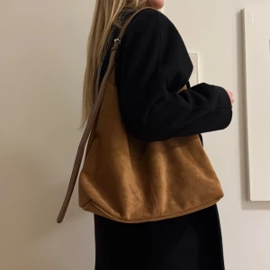 Brun mocka axelväska med justerbart band - Snygg axelväska i brun mocka med mjuk, slouchy form och justerbart axelband i matchande brunt. Väskan är rymlig och har en minimalistisk design utan synliga detaljer eller mönster. Perfekt för dig som gillar en stilren och trendig look.