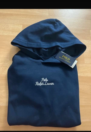 Mörkblå hoodie från Polo Ralph Lauren - Snygg mörkblå hoodie från Polo Ralph Lauren med vit broderad logga på bröstet och ärmen. Klassisk huva och stor magficka framtill. Mjuk bomullskvalitet och normal passform, perfekt för chill dagar. Stilren design med diskreta detaljer.