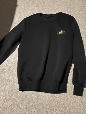 Svart sweatshirt från Stone Island - Svart sweatshirt från Stone Island med klassisk rund hals och ikonisk logga på bröstet. Tröjan har ribbade muddar vid ärmslut och midja. Perfekt för dig som gillar stilrena och snygga streetwear-plagg.pros diskuteras 