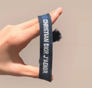 Mörkblå Dior J'Adior armband - Snyggt mörkblått tygarmband från Christian Dior med vit broderad text 'CHRISTIAN DIOR J'ADIOR'. Armbandet har flätad knytning och tofsar i ändarna, perfekt för att ge din outfit en lyxig touch.