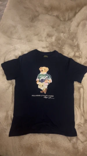 Mörkblå Polo Bear t-shirt Ralph Lauren - Mörkblå t-shirt från Ralph Lauren med ikoniska Polo Bear-trycket på bröstet. Klassisk rund hals och korta ärmar.                                                                   Säljer den för att den inte passar mig