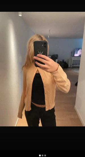 Aprikos stickad  kofta med knappar - Snygg aprikos kofta med rund hals och knappar hela vägen framtill. Modellen är klassisk med ribbade muddar vid ärmslut och nederkant. Perfekt att slänga över en topp för en chill och fräsch look. Materialet känns mjukt och lätt, passar dig som gillar enkel stil.