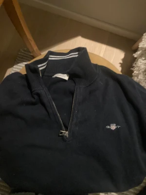 Mörkblå half zip tröja från GANT - Snygg mörkblå tröja från GANT med half zip och broderad logga på bröstet. Kragen har vita ränder som ger en sportig vibe. Perfekt för dig som gillar klassisk stil med en twist. Materialet känns mjukt och är troligtvis bomull.