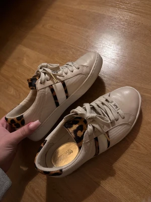 Beige sneakers från Michael Kors - Snygga beige sneakers från Michael Kors med leoparddetaljer på häl och sidor. Skorna har snörning, platt sula och rund tå. Materialmix av skinn och syntet, med vita och leopardmönstrade ränder på sidan. 