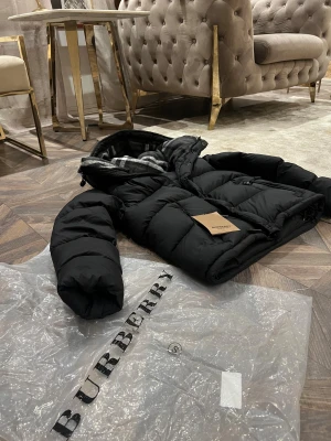 Svart pufferjacka från Burberry - Säljer en svart pufferjacka från Burberry med stor logotyp på bröstet och klassiskt rutigt foder i huvan. Jackan har dragkedja, flera fickor och en riktigt fluffig känsla. Perfekt för kalla dagar när du vill vara både varm och snygg.