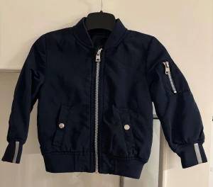 Mörkblå Skill bomberjacka med dragkedjor - Snygg mörkblå bomberjacka med silvriga dragkedjor, ribbade muddar och fickor med tryckknappar. Jackan har en klassisk bomberkrage och en extra ficka med dragkedja på ärmen. Perfekt för dig som gillar stilren och sportig look.