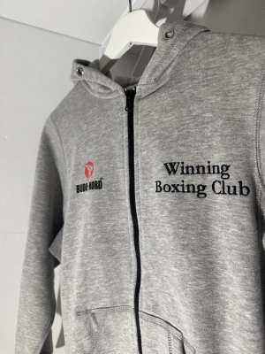 Grå hoodie Budo-Nord Winning Boxing Club - Grå hoodie från Budo-Nord med broderad text 'Winning Boxing Club' fram och bak. Tröjan har huva med tryckknappar, hel dragkedja och två fickor framtill. Perfekt för dig som gillar boxning eller vill ha en sportig look.
