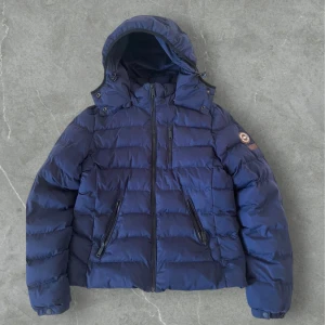 Superdry dunjacka   - Säljer en mörkblå dunjacka från Superdry med avtagbar huva och dragkedjor på fickorna. Jackan har ett broderat Superdry-märke på ärmen och är vadderad för extra värme. Perfekt för kalla dagar.